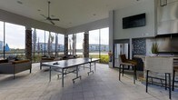 The Sun Kitchen is a single-story, semi-open recreation and lounge space designed for casual gathering and leisure. The area is defined by its expansive floor-to-ceiling windows and open-air wall sections that flood the interior with natural light while offering unobstructed views of the waterfront and neighboring community buildings. Stone-clad columns punctuate the perimeter, adding a grounded, architectural character to the otherwise airy environment. The ceiling is elevated, contributing to a spacious, breathable atmosphere, and a large ceiling fan ensures comfortable airflow throughout. Underfoot, large-format tile flooring in a neutral stone finish lends a clean, durable surface well-suited to an active recreational setting. A full-size ping-pong table serves as the centerpiece of the space, encouraging active engagement. Along one wall, a stainless steel refrigerator and bar-height seating provide convenient amenities for socializing, while a wall-mounted flat-screen television adds an entertainment focal point. Lounge seating positioned near the open walls allows occupants to relax while taking in the outdoor scenery, making this space ideal for both recreation and unwinding.