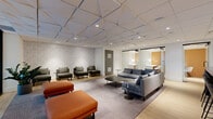This thoughtfully designed coworking space in California offers a sophisticated and functional environment spread across a single floor. The open-plan layout is anchored by a comfortable lounge area featuring a large sectional sofa in soft grey, complemented by olive-toned armchairs with ottomans and vibrant rust-orange benches, all arranged atop patterned area rugs over light hardwood flooring. A standout architectural feature is the geometric acoustic ceiling tile system, which adds both visual texture and sound management throughout the space. A sleek bar-height counter with a dark terrazzo surface serves as a central hub, lined with contemporary black barstools and illuminated by pendant lighting above. Glass-partitioned private offices or focus rooms line one side of the space, offering semi-private workspaces with warm amber accent walls visible through the transparent enclosures. Black-framed floor-to-ceiling glass walls and sliding doors create a clean, modern aesthetic while maintaining an open and airy feel. Recessed lighting throughout ensures the space is well-lit, while textured wallcoverings and curated greenery add warmth and character. The overall ambiance balances professional functionality with inviting, lounge-like comfort.