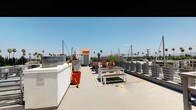 This rooftop terrace offers a generously sized open-air deck with sweeping panoramic views of the surrounding urban landscape, characterized by rows of tall palm trees set against a clear blue sky. The deck is finished in smooth concrete and is enclosed by contemporary metal railing panels that provide both safety and an unobstructed sightline to the horizon.

A central seating area anchors the space, featuring a white-framed outdoor dining table paired with matching benches and wood-slatted accents, set atop a striped outdoor rug. Potted plants and colorful foliage add greenery and visual interest throughout the terrace, softening the industrial character of the space.

On one side of the deck, a white stucco mechanical housing structure provides architectural contrast, while a rooftop access enclosure with a distinctive orange-accented door serves as the primary entry point to the terrace. Cable wire infrastructure is also present overhead, adding to the utilitarian yet functional design.

The terrace benefits from abundant natural sunlight throughout the day, making it a versatile outdoor space well-suited for gatherings, relaxation, or enjoying the open-sky surroundings.