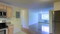 This single-floor space features a straightforward, functional layout with clean lines and neutral wall tones throughout. The living area is anchored by polished hardwood floors that reflect natural light streaming in through a window dressed with blue curtains, offering a view of greenery outside. An in-wall air conditioning unit and a baseboard heater provide year-round climate control. The open layout connects the entry and living zones through a wide archway, creating a seamless flow between spaces. An electrical panel is conveniently mounted on the wall near the entry, alongside an intercom system, indicating the practical utility features built into the design. The ceilings are standard height, contributing to the comfortable, manageable scale of the space. White baseboards frame the hardwood floors throughout, adding a clean, crisp finish. The overall ambiance is bright and neutral, providing a blank canvas well-suited for various furnishing and decorating styles. The unit's simple, uncluttered design emphasizes functionality while maintaining a welcoming atmosphere suitable for everyday living.