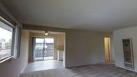 This single-floor unit presents an open-concept layout that connects the main living area to a secondary space, creating a seamless flow between rooms. The living area is finished with wall-to-wall carpeting and neutral-toned walls, offering a clean, move-in-ready canvas. A ceiling fan with a light fixture is centrally positioned in the inner room, providing both ventilation and ambient lighting.

A sliding glass door opens to a private balcony, which overlooks a neighboring structure and features a wooden railing. This access point allows natural light to filter generously into the interior, brightening the tile-floored transitional zone between the living space and the kitchen or utility area. Upper cabinetry is visible in this section, along with a side-by-side washer and dryer unit, indicating in-unit laundry convenience.

The main living space features smooth, neutral-painted walls and plush carpeting underfoot, contributing to a quiet and comfortable atmosphere. A doorway visible on the right side suggests additional rooms beyond the main living area. Overall, the space offers a functional and straightforward layout suited for everyday living.