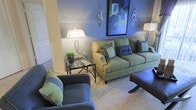 This single-floor living space showcases a thoughtfully designed interior centered around a cohesive blue and green color palette. The living area serves as the focal point, featuring a sage green sofa adorned with an assortment of decorative pillows in teal, navy, and patterned fabrics, complementing a matching armchair that adds depth to the seating arrangement. A dark leather ottoman provides both functionality and contrast within the space.

The walls are painted in a calming periwinkle-blue tone, accented by a horizontal stripe detail that adds visual interest and architectural dimension. A bold abstract artwork in blues and greens anchors the main wall, flanked by elegant silver ring-shaped wall sconces that enhance the refined aesthetic.

A glass-top side table with a sleek metal frame holds a classic table lamp and small potted greenery, contributing to the layered, lived-in feel of the room. A neutral area rug grounds the seating arrangement, while a structural column adds an architectural element to the layout. Ambient and accent lighting work together to create a warm, welcoming atmosphere throughout the space.