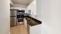 Located at 1107 Garner Rd in Raleigh, NC, this single-floor unit offers a clean, modern aesthetic throughout. The living area is finished with light wood-look flooring and neutral warm-gray walls, creating a bright and welcoming atmosphere. A large sliding glass door fitted with vertical blinds allows generous natural light to fill the space and provides convenient outdoor access.

The kitchen is a standout feature, showcasing white shaker-style cabinetry, dark granite countertops, and a full suite of stainless steel appliances including a refrigerator, electric range, over-the-range microwave, and dishwasher. A stainless steel undermount sink with a modern faucet completes this functional and stylish cooking space.

The bathroom features a full tub-shower combination lined with crisp white tile, offering a clean and practical design. The unit also includes a dedicated walk-in closet outfitted with built-in shelving and a hanging rod, providing ample organized storage.

The dollhouse view reveals a compact yet efficiently laid-out floor plan where the living, kitchen, bathroom, and closet areas are thoughtfully arranged. Consistent light flooring and white trim flow throughout, lending the entire unit a cohesive and polished look. The overall ambiance is bright, neutral, and move-in ready.