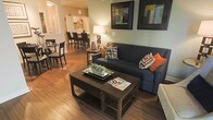 This single-floor residence offers an open-concept layout that effortlessly connects the living, dining, and kitchen areas, creating a cohesive and functional living environment. The living area serves as the heart of the space, anchored by rich hardwood flooring that extends throughout. A navy blue sofa accented with vibrant orange and geometric-patterned throw pillows adds a pop of color and personality, complemented by a dark wood coffee table that grounds the seating arrangement. Framed artwork adorns the sage-green accent walls, lending a curated, finished aesthetic. A warm table lamp provides inviting ambient lighting, enhancing the cozy yet modern atmosphere. The dining area transitions naturally from the living space, featuring a round table paired with sleek dark chairs, ideal for intimate gatherings. Beyond the dining area, the kitchen is bright and well-lit, showcasing bar-height seating along a counter, white cabinetry, and ample workspace. The open sightlines between all three areas promote a sense of spaciousness, making the layout feel airy and connected. The consistent use of dark wood tones in the furnishings and flooring ties the entire space together with a warm, contemporary elegance.