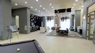 This expansive single-floor lounge is designed with a sleek, contemporary aesthetic that blends functionality with upscale style. The open-plan layout seamlessly connects several distinct zones, creating a versatile space suited for both social gatherings and relaxed lounging. Large windows dressed with floor-length curtains flood the interior with natural light, complementing the neutral palette of light tile flooring and gray walls.

At the forefront is a billiards table, anchoring the recreational area with a bold, game-room feel. Beyond it, multiple seating arrangements are thoughtfully positioned throughout the space, including plush armchairs and sofa groupings set atop a geometric-patterned area rug. A dramatic chandelier with dark, cascading elements serves as a striking focal point over one of the lounge sections, adding a touch of glamour.

A built-in bar with dark cabinetry and backlit shelving runs along one wall, accompanied by a row of sleek bar-height seating—ideal for socializing. Recessed lighting across the ceiling provides consistent, well-distributed illumination throughout. The overall ambiance is modern and refined, offering an inviting yet polished environment.