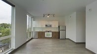 This single-floor space showcases a clean, contemporary design centered around an open-concept layout. The kitchen runs along one wall in a well-organized galley configuration, featuring a thoughtful combination of crisp white upper cabinets and warm wood-toned lower cabinetry. Stainless steel appliances—including a refrigerator, dishwasher, and built-in microwave—complement the white gas range, creating a functional and visually cohesive cooking area. A light green glass backsplash adds a subtle pop of color to the otherwise neutral palette.

Track lighting above the kitchen provides focused illumination, while the light-toned laminate hardwood flooring extends throughout the open living area, adding warmth and continuity to the space. A standout feature is the floor-to-ceiling window wall along the left side, which allows abundant natural light to pour in and offers views of surrounding greenery. The white walls and pale ceiling enhance the airy, open feel of the unit. The overall ambiance is modern and minimalist, appealing to those who value clean lines and functional design.