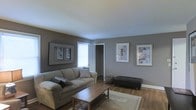 This single-story space centers around a well-appointed living room that combines functionality with understated style. The room is finished with warm hardwood flooring throughout, which contrasts attractively against the cool, medium-grey painted walls. A light-toned ceiling enhances the sense of openness in the space.

The living area is furnished with a comfortable, beige upholstered sofa accented with decorative pillows, paired with a wooden coffee table featuring an X-base design. A dark rectangular ottoman provides additional seating or surface space, while a neutral area rug anchors the seating arrangement. Three framed artworks are displayed across the walls, adding visual interest and a curated touch to the room's décor.

Natural light enters through a window near the entryway, brightening the space during the day. A doorway visible from the living area leads to an adjoining room, suggesting additional functional spaces beyond. A small woven basket adds a subtle decorative accent near the seating area. Overall, the space presents a clean, cohesive aesthetic with a neutral color palette that feels both practical and inviting.
