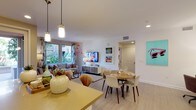 This single-story residence showcases a bright, open-plan layout that integrates the kitchen, dining, and living areas into one cohesive and visually dynamic space. The kitchen features a bold yellow island countertop with stylish pendant lighting overhead, providing both functional workspace and an eye-catching focal point. Light wood-tone flooring flows continuously throughout, lending a sense of warmth and visual continuity across the space.

The dining area is positioned centrally, anchored by a round wooden table surrounded by a mix of modern white and wood-toned chairs, creating an inviting setting for gatherings. Framed artwork and a colorful flamingo-themed wall mural introduce playful, artistic character throughout the space.

The living area is well-lit by large windows that invite natural light and offer views of lush greenery outside. A wall-mounted flat-screen television, a media console, and a comfortable sofa with decorative pillows complete the lounge setting. Recessed lighting and clean white walls contribute to the bright, airy atmosphere. The overall ambiance balances contemporary design with a lively, artistic personality.