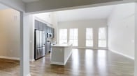 This contemporary 2-floor residence offers a thoughtfully designed open-concept layout that seamlessly connects the kitchen and living areas. The kitchen is a standout feature, equipped with stainless steel appliances including a full-size refrigerator, microwave, and range. Sleek gray cabinetry provides ample storage, while a large central island with a white countertop and integrated sink serves as both a functional workspace and a natural gathering point. The open living area is bathed in natural light, thanks to a row of large windows dressed with white blinds that frame views of the outdoors. Soaring ceilings amplify the sense of space, creating an airy and expansive atmosphere throughout. Warm hardwood-style flooring runs continuously across the main level, tying the spaces together with a cohesive and polished look. Neutral wall tones in soft gray and white complement the modern finishes, lending the interior a clean, versatile aesthetic. The two-floor layout offers additional living or sleeping space above, making this a well-rounded and functional home suited for a variety of lifestyles.