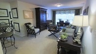 Located at 1201 Aragon Dr in Knightdale, NC, this single-floor residence offers a thoughtfully designed living space that blends classic elegance with everyday comfort. The living area serves as the heart of the home, showcasing rich, dark wood furnishings including a stately armoire and a refined coffee table that anchor the room with a sense of sophistication. Plush, neutral-toned carpeting runs underfoot, providing a warm and inviting foundation throughout. Seating is arranged to encourage both relaxation and conversation, with patterned accent chairs and a striped sofa complemented by coordinating throw pillows in deep greens and blacks. A large window dressed with decorative valances allows natural light to fill the room, brightening the soft, neutral wall tones and creating an airy ambiance. A dedicated desk area near the window provides a functional workspace integrated seamlessly into the living environment. Decorative accents, including framed artwork and curated tabletop arrangements, add personality and polish to the space. The overall atmosphere is warm, classic, and carefully curated, reflecting a home designed with both style and livability in mind.