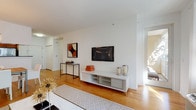 This well-appointed single-floor residence offers a thoughtfully designed layout that balances open communal spaces with comfortable private retreats. The living room serves as the heart of the home, showcasing warm hardwood flooring, crisp white walls, and a generous arrangement of large windows dressed with sheer curtains that invite an abundance of natural light and views of lush greenery outside. A wall-mounted flat-screen television and a sleek media console anchor the entertainment area, while a pass-through opening connects the space to the kitchen beyond.

The dining area flows naturally from the living room and features hardwood flooring, a stylish drum pendant light, and an arched mirror that adds depth and elegance to the room. Adjacent, the galley kitchen is equipped with warm wood cabinetry, dark countertops, tile flooring, and stainless steel appliances including a refrigerator and dishwasher.

The two bedrooms offer a serene retreat, each carpeted for comfort and filled with soft natural light from well-placed windows. One bedroom features a neutral accent wall with decorative touches, while both offer ample closet storage.

The full bathroom is finished with tile flooring, a warm wood vanity, and clean white fixtures, completing this cohesive and inviting residence.