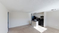 This single-floor space features an open-concept design that seamlessly connects the living area to the kitchen. The living area is finished with wall-to-wall beige carpet, creating a warm and neutral foundation, while the textured popcorn ceiling adds a classic touch overhead. Natural light filters in through a window, brightening the space noticeably.

The kitchen is positioned toward the rear and is equipped with black appliances, including a refrigerator, gas range with overhead exhaust hood, and a microwave. White upper cabinetry provides ample storage, contrasting cleanly against the dark countertops. A peninsula with a dark countertop extends into the transitional space, offering additional prep area and a natural visual divider between the kitchen and living zone. The kitchen features tile or vinyl flooring, distinct from the carpeted living area.

The overall palette is neutral and understated, with white walls and ceilings contributing to a clean, functional aesthetic. The layout is practical and efficient, making effective use of the available space while maintaining an open and airy feel throughout.