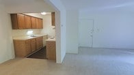 This single-floor residence features two bedrooms and one bathroom within a well-utilized floor plan. The living area is finished with neutral wall-to-wall carpeting, creating a clean, cohesive atmosphere throughout the main space. A front entry door opens directly into the living area, providing straightforward access to the home.

The kitchen is accessible via an arched passthrough opening, which visually connects it to the living space while maintaining a distinct functional zone. The kitchen features warm-toned wood cabinetry providing ample storage, a white electric range, a dishwasher, and a sink positioned along the counter. A peninsula with a raised bar edge extends from the cabinetry, offering additional workspace and a casual dining option. Dark flooring in the kitchen contrasts with the carpeted living area, clearly delineating the two spaces.

The overall aesthetic is straightforward and practical, with white walls and warm wood tones contributing to a neutral interior palette. The layout is designed to maximize usability, making efficient use of the available square footage across the living, dining, and kitchen areas.