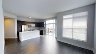This single-floor space showcases a thoughtfully designed open-concept layout that seamlessly integrates the kitchen and living areas. The kitchen is outfitted with dark, espresso-toned cabinetry that contrasts sharply against a bright white island countertop, creating a bold visual centerpiece. Stainless steel appliances, including a refrigerator and built-in microwave, complement the contemporary aesthetic, while light-toned tile backsplash adds a subtle texture to the cooking area. Pendant lighting above the island enhances both functionality and style.

The living area extends generously from the kitchen, unified by wide-plank hardwood-style flooring in a warm gray tone that runs throughout the space. Natural light floods the interior through large floor-to-ceiling sliding glass doors that lead to an outdoor balcony, offering views of the surrounding landscape. An additional oversized window fitted with horizontal blinds further brightens the living area. The neutral gray walls and clean white trim maintain a crisp, modern ambiance throughout, making the space feel open, airy, and versatile.