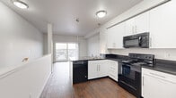 Located at 5601 Gordon Rd in Regina, Saskatchewan, this efficiently designed unit offers a single-floor open-concept layout that combines a kitchen and living area into one cohesive living space. The kitchen is well-appointed with crisp white cabinetry, dark countertops, and a full suite of black appliances including a refrigerator, electric range, over-the-range microwave, and dishwasher. Pendant lighting and flush-mount ceiling fixtures illuminate the space, while rich dark hardwood-style flooring runs throughout, adding warmth and visual continuity. The kitchen flows naturally into the living area, which is bright and airy thanks to a large sliding glass door that leads to a private balcony, offering open views of the surrounding Regina neighbourhood. Natural light pours into the living space, enhancing the clean, neutral palette of white walls and grey tones that define the unit's modern aesthetic. The entryway is tidy and functional, featuring a closet for storage and clean white panel doors. The overall design is contemporary and understated, making efficient use of the available space while maintaining a comfortable and inviting atmosphere suited to everyday living.