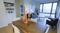 This single-floor space showcases a thoughtfully designed open-concept layout that seamlessly connects the dining area, living room, and bedroom. The dining area features a substantial rustic wood table paired with white chairs, centered with decorative terracotta vases that add a warm, artistic touch. Adjacent to the dining space, the living room is furnished with a navy blue sofa accented by patterned throw pillows, a round marble-top coffee table, and a textured area rug that defines the seating zone. A wall-mounted mini-split HVAC unit ensures year-round climate control. Large floor-to-ceiling windows flood the living area with natural light and offer expansive urban views, creating a bright and airy atmosphere. An adjustable arc floor lamp provides functional and ambient lighting throughout the shared space. Through an open doorway, the bedroom is visible, featuring a blue accent wall that adds depth and character. The overall design balances modern furnishings with warm, natural textures, resulting in a cohesive and inviting living environment.