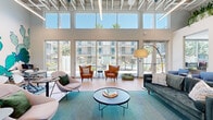Located at 5300 E Washington St in Phoenix, AZ, this single-floor communal lounge space offers a generously sized, open-plan interior designed with a modern aesthetic. The architectural design is defined by soaring double-height ceilings with exposed structural elements and a coffered ceiling system, lending the space an airy, industrial-contemporary character. Abundant natural light floods the interior through expansive floor-to-ceiling glass walls and large sliding doors, providing direct views of an outdoor pool area framed by palm trees.

The main lounge area features warm hardwood-style flooring throughout, anchored by vibrant teal-patterned area rugs. Seating arrangements include a mix of tufted sofas, mid-century modern accent chairs in cognac leather and soft gray upholstery, and bistro-style tables. A bold desert-themed mural with cactus motifs serves as a striking focal point on one wall, complemented by curated artwork and lush hanging greenery integrated into elevated ceiling planters.

A smaller seating nook near the glass entrance provides an intimate retreat with direct outdoor views, while a kitchenette area with light wood cabinetry and a utility sink supports functional use. Private office or manager spaces are also accessible within the layout, rounding out this well-appointed, amenity-rich communal environment.