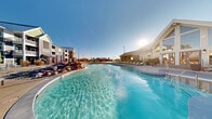 Located at Bear Run Point in Colorado Springs, CO, this resort-style pool amenity complex is designed for year-round enjoyment and offers an impressive array of outdoor and semi-outdoor features. The centerpiece is a generously sized swimming pool with crystal-clear water, complemented by a separate hot tub spa, both framed by decorative boulder landscaping and wide concrete pool decking lined with lounge chairs.

Adjacent to the pool, a covered cabana area serves as an inviting gathering space. It features a long granite-topped bar with barstool seating, pendant lighting suspended from an exposed beam ceiling, and an outdoor television — ideal for social events. A second covered patio area with a vaulted, wood-planked ceiling offers round dining tables with chairs and an additional television behind sliding barn-style doors, connecting directly to the interior clubhouse via French doors.

The clubhouse building itself is architecturally striking, featuring soaring gabled glass windows, a stone chimney, and a welcoming walkway with xeriscape landscaping suited to Colorado's climate. The surrounding grounds include open concrete patio areas with umbrella-shaded dining sets, thoughtfully arranged greenery, and views of the multi-story residential buildings that frame the community. The overall atmosphere is polished, resort-inspired, and well-suited for both relaxation and community gatherings in all seasons.