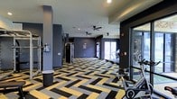 This fitness center delivers a dynamic and motivating environment for exercise, thoughtfully designed with both function and aesthetics in mind. The space is anchored by a striking geometric-patterned carpet in bold yellow, grey, and black tones, adding a high-energy visual character to the floor throughout the room. Deep charcoal-grey walls create a sleek, contemporary backdrop that complements the modern design scheme.

The facility is generously sized, offering ample open floor space to accommodate a diverse range of workout activities. Ceiling fans are mounted overhead to promote air circulation during intensive training sessions, while recessed lighting ensures the space is consistently and evenly lit. Large glass windows and doors line one side of the room, flooding the interior with natural light and providing views to an adjacent outdoor area, enhancing the overall sense of openness.

The equipment layout is well-organized, featuring cardio machines — including treadmills visible along the windowed wall — as well as strength training equipment and a rowing machine positioned toward the front. A cable resistance machine adds to the versatility of the strength training offerings. The combination of natural light, bold design choices, and comprehensive equipment makes this fitness center a functional and visually engaging space suited for a full-body workout routine.