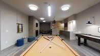 This single-floor amenity lounge offers a well-rounded recreational space designed for socializing and leisure. At the center of the room sits a full-size pool table, which serves as the focal point and is complemented by ample surrounding space for comfortable gameplay. The room's design blends modern and industrial aesthetics, highlighted by exposed brick columns and walls that add warmth and texture throughout.

Toward the back of the space, a cozy seating area with chairs is anchored by a fireplace, creating an inviting retreat for relaxation. A wall-mounted television is positioned nearby, adding an entertainment option to this lounge corner. Blue upholstered ottomans provide additional casual seating options near the pool table.

A bar or kitchenette area is situated to one side, featuring a countertop, cabinetry, and what appears to be a sink, making the space well-suited for hosting. Overhead, a combination of flush-mount and pendant lighting fixtures illuminates the room effectively, including a statement pendant light above the bar area. Hard flooring runs throughout, contributing to the space's clean, contemporary feel.