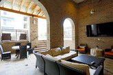 This stylish amenity lounge blends rustic and contemporary design elements to create a refined communal space. The architecture is defined by striking exposed brick walls and elegant arched openings that frame views of an outdoor courtyard, establishing a seamless connection between interior and exterior areas. Stone tile flooring in a geometric pattern runs throughout, adding texture and visual interest underfoot.

The lounge is furnished with cream-toned sectional and lounge seating arranged to encourage relaxed gathering and social interaction. Wall-mounted flat-screen televisions are positioned throughout, catering to entertainment needs across different seating areas. Pendant lighting and recessed fixtures illuminate the space, complementing the warm tones of the brick and tile.

Large black-framed glass windows and doors offer views into adjacent interior spaces, including a staircase with hardwood flooring and a game room featuring a billiards table — hinting at a broader suite of amenities within the building. The soaring brick walls and open archways lend a generous sense of height and openness to the lounge. The outdoor courtyard, accessible via wide stepped entries with metal railings, extends the usable gathering space and enhances the overall ambiance of this well-appointed communal area.
