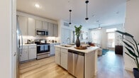 This single-floor residence is designed with a clean, contemporary aesthetic that balances functionality and style. The open-concept kitchen serves as the heart of the home, featuring a central island with an undermount sink, stainless steel appliances including a side-by-side refrigerator, electric range, microwave, and dishwasher, all complemented by soft gray cabinetry, white quartz countertops, and a subway tile backsplash. Pendant lighting above the island adds a polished touch, while light wood-style flooring extends seamlessly into the adjoining living area, which benefits from abundant natural light through multiple windows fitted with blinds and sheer curtains.

The living room offers a comfortable, well-lit retreat, while a dedicated office space provides a functional work area with carpeted flooring, large windows with outdoor views, and ample room for a substantial desk setup. The bedrooms are carpeted and bright, each with generous window exposure and ceiling fans. Two bathrooms are finished with light wood flooring, gray vanity cabinetry, white quartz countertops, and large mirrors. The primary bathroom features a dual-sink vanity and a combination tub and shower, while the secondary bathroom offers a mirrored closet and direct bedroom access. The overall ambiance is bright, neutral, and cohesive throughout.