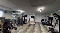 Located at 850 Imperial Blvd in Sugar Land, Texas, this fitness center is a thoughtfully designed, single-floor facility offering a comprehensive range of workout spaces and equipment. The main exercise floor is finished with a distinctive hexagonal-patterned carpet in shades of green, gray, and white, lending the space a modern, energetic aesthetic. The area is generously outfitted with professional-grade Cybex cardio equipment, including treadmills, ellipticals, and stationary bikes, as well as a variety of strength training machines such as leg press, cable systems, and multi-functional units. Full-length wall mirrors expand the sense of space and allow users to monitor their form throughout their workout. Recessed fluorescent lighting ensures consistent, bright illumination across the floor.

A dedicated group exercise studio is separated from the main gym floor by floor-to-ceiling glass walls, providing visual openness while maintaining acoustic separation. This studio features smooth hardwood-style flooring, a large wall-mounted display screen, ambient cove lighting with warm tones along the ceiling beams, and a built-in sound system with ceiling-mounted speakers. A mirrored accent wall further enhances the room's spaciousness. Louver-paneled doors provide access to additional utility and storage areas. Natural light filters in through windows, contributing to an inviting and motivating environment throughout the facility.