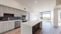 This contemporary single-floor residence showcases a clean, modern aesthetic throughout. The open-concept kitchen serves as a central hub, outfitted with flat-front cabinetry in a warm gray wood-grain finish, a textured tile backsplash, and a full suite of stainless steel appliances including a gas range, dishwasher, and French door refrigerator. A large center island with a crisp white quartz countertop and dark base offers additional prep space and casual seating. Light hardwood flooring runs through the main living areas, which are anchored by expansive floor-to-ceiling windows framed in bold black that invite abundant natural light and offer sweeping treetop and cityscape views. The living area is generously proportioned with recessed lighting above. Bedrooms feature plush carpet, clean white walls, and large windows continuing the bright, airy feel. The full bathroom is elegantly appointed with a dual-sink vanity featuring dark cabinetry, a white quartz countertop, a full-length mirror, and a soaking tub with subway tile surround, complemented by large-format floor tiles and a window overlooking the outdoors.