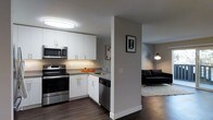 This single-floor space showcases a thoughtfully designed open-concept layout that seamlessly connects the kitchen and living area. The kitchen is outfitted with clean white cabinetry featuring brushed metal hardware, offering generous storage across both upper and lower cabinets. Stainless steel appliances — including an electric range, built-in microwave, and dishwasher — lend a modern, functional aesthetic, while the countertops provide ample workspace. A stainless steel sink is positioned beneath under-cabinet lighting that brightens the workspace effectively. Rich, dark hardwood flooring runs throughout the space, providing a warm contrast to the crisp white and grey palette. A structural half-wall with a column detail serves as a subtle visual divider between the kitchen and the adjacent living area, maintaining an open feel while defining each zone. The living area features a cozy layout anchored by a plush dark sofa set atop a light area rug, with ambient floor lamp lighting creating a welcoming atmosphere. Neutral grey walls and flush ceiling lighting complete the interior, giving the space a clean, contemporary character that is both practical and inviting.