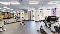 This fitness center at Riverwalk in Plainsboro, NJ, offers a thoughtfully designed, single-floor exercise facility built to support a wide range of workout routines. The space is defined by durable speckled rubber flooring throughout, a drop ceiling with recessed panel lighting, and large floor-to-ceiling mirrors that enhance the sense of openness and allow users to monitor their form.

The cardio zone is a standout feature, lined with a row of treadmills and ellipticals positioned in front of a warm, rustic wood-paneled accent wall. Multiple flat-screen televisions are mounted along this wall, providing entertainment during workouts. Adjacent to the cardio equipment, a dedicated strength and functional training area houses a selection of free weights, resistance machines, stability balls, and benches, keeping all essential equipment easily accessible.

A vibrant, large-scale motivational mural—featuring bold geometric patterns in green, blue, and yellow hues with phrases such as "In It for the Long Run" and "You Can and You Will"—adds energy to the space. A water fountain station and clearly marked restrooms are conveniently located nearby. Glass doors along one wall provide views of the outdoors and allow natural light to supplement the interior lighting, contributing to a bright, welcoming atmosphere.