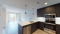 This contemporary single-floor residence is thoughtfully designed with a clean, modern aesthetic throughout. The open-concept kitchen serves as a focal point of the living space, featuring dark espresso-toned cabinetry, crisp white quartz countertops, and a peninsula island with an undermount sink. Stainless steel appliances — including a range, microwave, and dishwasher — complement the kitchen's polished look, while two stylish pendant lights with a brushed brass finish add warmth overhead. Light wood-tone flooring flows throughout the main living areas, enhancing the bright and airy atmosphere. Large sliding glass doors in the living area allow abundant natural light to pour in and provide a seamless connection to the outdoors. The bedroom is carpeted for comfort and features its own sliding glass door opening to a private balcony, offering pleasant outdoor views. An in-unit washer and dryer provide added convenience. The bathroom is outfitted with a wide vanity topped with white countertops, dark wood-grain cabinetry, a large mirror, and ample storage. The overall design is characterized by a neutral color palette, clean lines, and contemporary finishes that create a cohesive and functional living environment.