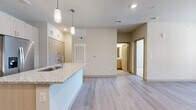 This thoughtfully designed one-bedroom, one-bathroom residence offers a comfortable and functional single-floor layout. The open-concept living and kitchen area is anchored by a granite-topped island with a built-in sink and pendant lighting overhead, creating an inviting focal point. The kitchen is equipped with stainless steel appliances, including a French door refrigerator, electric range, and over-the-range microwave, complemented by light wood cabinetry and a classic subway tile backsplash. Light-toned luxury vinyl plank flooring runs throughout the main living areas, adding warmth and continuity. The living room features a ceiling fan, natural light from multiple windows, and a glass-panel entry door that enhances brightness. The bedroom is carpeted for comfort, includes its own ceiling fan, and benefits from natural light through a window with blinds. A dedicated walk-in closet offers built-in shelving and hanging rods, providing ample organization. The bathroom is finished with a granite dual-sink vanity, light wood cabinetry, a soaking tub with large-format tile surround, and durable plank-style flooring. Neutral, warm tones throughout lend the space a clean, cohesive aesthetic.