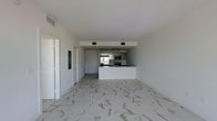 This single-floor space showcases a clean, contemporary design centered around an open-concept layout that connects the living area to the kitchen. The flooring throughout is a striking marble-look porcelain tile in white and grey tones, lending an elegant and upscale feel to the interior. The living area is generously proportioned, offering a bright and airy atmosphere thanks to the crisp white walls and smooth white ceiling.

The kitchen is positioned toward the rear of the space and features a peninsula-style counter that provides both workspace and a natural divider between the cooking and living zones. It is equipped with stainless steel appliances, including a refrigerator, microwave, and range, alongside white cabinetry and dark countertops that create a modern contrast. Track lighting above the kitchen enhances visibility and adds a functional design element. Ceiling vents are evenly distributed, indicating a central HVAC system. A doorway and entry panel are visible along the side wall, contributing to the unit's practical layout. Overall, the space presents a minimalist, move-in-ready aesthetic.