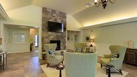 This single-floor space exudes a warm and inviting ambiance, thoughtfully designed for comfort and relaxation. The centerpiece of the main lounge area is a striking floor-to-ceiling stone fireplace, crafted from natural stacked stone that adds rustic character and visual drama to the room. A flat-screen television is mounted above the fireplace, making the space both functional and cozy. The vaulted ceilings lend an airy, open feel, while soft, recessed and accent lighting creates a welcoming glow throughout. The seating arrangement features a curated selection of sage green wingback chairs and a complementary curved sofa, all positioned to encourage conversation and relaxation. Decorative throw pillows in coordinating blue and white tones add subtle style accents. The flooring consists of neutral-toned tiles that flow seamlessly throughout the space. Wainscoting and clean wall paneling details contribute a classic, refined touch to the interior design. A traditional chandelier and table lamps provide layered lighting that enhances the room's warm palette. Framed artwork and thoughtful décor accessories complete the polished yet comfortable aesthetic, making this a well-appointed communal or gathering space.