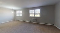 This single-floor unit offers a practical and straightforward layout, as revealed by its 3D floor plan, which shows a spacious living area, a separate bedroom, a kitchen, and a bathroom arranged in a functional configuration. Throughout the carpeted rooms, neutral gray walls and white trim create a clean, understated aesthetic.

The expansive main living area is one of the most notable spaces, featuring multiple white-framed windows that allow generous natural light to fill the room. A wall-mounted air conditioning unit and baseboard heating run along the exterior wall, providing year-round climate control. An additional carpeted bedroom offers similar features, including a window with outdoor views, baseboard heating, and a wall A/C unit. This room also includes a sliding-door closet with a hanging rod, providing practical storage.

The kitchen is equipped with warm-toned wood cabinetry, dark granite-style countertops, and black appliances including a gas range, built-in microwave, dishwasher, and full-size refrigerator. Dark stone-look tile floors add durability to this functional space. The bathroom features the same tile flooring, complemented by large-format gray tile surrounding a full bathtub and shower combination.