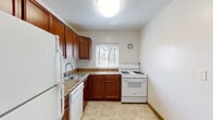This single-floor residence offers a well-organized layout with distinct living, sleeping, and utility areas. The kitchen is equipped with rich, warm-toned wood cabinetry, stone-patterned laminate countertops, a stainless steel sink, and white appliances including a gas range, dishwasher, and refrigerator. A window above the counter allows natural light to brighten the workspace, while light tile flooring completes the practical design. The open living area features neutral carpet flooring and white walls, creating a clean, versatile canvas. A hallway connects the main living space to the rest of the unit, with the refrigerator and additional rooms visible from this central corridor. The bedrooms are carpeted and each includes a ceiling fan with light fixture, a window with blinds offering natural light and outdoor views, and a wall-mounted heating unit. Closet storage is well-represented throughout, with both louvered bi-fold and standard swing-door closets providing ample hanging and shelving space. The bathroom features distinctive pink ceramic tile lining the tub surround and lower walls, a white soaking tub, and a window for natural ventilation. Overall, the unit presents a functional and straightforward residential layout with consistent finishes throughout.