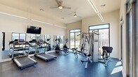 This fitness center in Denham Springs, Louisiana, offers a bright, modern workout environment designed to support a full range of exercise routines. The single-floor facility is bathed in natural light, thanks to expansive floor-to-ceiling windows along multiple walls that provide open views of the surrounding outdoor scenery. The high ceilings, fitted with ceiling fans and sleek linear LED lighting, contribute to an airy and comfortable atmosphere throughout.

The space is equipped with a comprehensive selection of cardio machines, including multiple treadmills with individual console displays and elliptical trainers, ideal for sustained aerobic workouts. Strength training is well-represented with a multi-station cable machine and various resistance equipment to accommodate a wide range of exercises. A large wall mirror spans one side of the room, enhancing the sense of space and allowing users to monitor their form during workouts.

The flooring consists of durable, speckled rubber gym flooring that provides both safety and comfort underfoot. A Coca-Cola vending machine is conveniently located near the entrance, offering easy refreshment access. A wall-mounted flat-screen television is positioned for viewing during cardio sessions. Additional doors provide access to utility and restroom facilities. The overall design is clean, functional, and welcoming, making it a practical and well-appointed fitness destination.