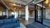 This single-floor space showcases a compelling industrial aesthetic defined by exposed ductwork, conduit, and structural beams running across the high ceiling, all illuminated by warm Edison-style pendant lighting. The open-concept layout offers generous square footage, creating a versatile and airy environment suited for a variety of uses.

The main floor features polished concrete flooring throughout, lending a clean yet rugged character to the space. Structural white-painted columns punctuate the floor plan, while a striking exposed brick wall serves as a dramatic focal point along one side, adding warmth and texture to the industrial design.

A lounge area near the entrance includes blue upholstered seating arranged on area rugs, providing an inviting and comfortable gathering spot. Further into the space, a counter or bar-height surface is visible alongside a railing system, suggesting a tiered layout or lower-level access point. Large windows and glass entry doors along the perimeter allow natural light to filter in, enhancing the open and welcoming atmosphere.

Overall, the space balances raw industrial elements with thoughtful design touches, creating a dynamic and functional environment.