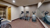 Located at 7700 Cahill Rd in Edina, Minnesota, this fitness center offers a practical and well-equipped space for exercise. The main workout area is carpeted in a durable, patterned commercial carpet and features neutral-toned walls with recessed lighting that provides consistent, even illumination throughout. The room is outfitted with two treadmills equipped with digital display consoles, an elliptical trainer, and an air resistance exercise bike, offering a well-rounded selection of cardio equipment. A large wall-mounted mirror spans one side of the room, visually expanding the space and allowing users to monitor their form during workouts. A large floor fan provides additional ventilation, complementing the room's functional design. An adjoining anteroom serves as an entry and transitional area, featuring a wood-panel door with an emergency call sign posted nearby, as well as storage for bicycles. A separate shower room is accessible from this area, finished with white subway tile walls, a tub-shower combination with a curtain rod, and a grab bar for safety, making it convenient for users to freshen up after a workout. Together, the rooms provide a complete and practical fitness facility.
