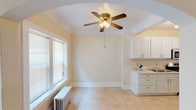 This single-floor space showcases a blend of classic architectural character and functional design throughout its rooms. Elegant arched doorways connect the living areas, adding a timeless charm to the overall layout. The spacious living room is a standout feature, highlighted by a traditional white-mantled fireplace, plush wall-to-wall carpeting, crown molding, and a ceiling fan with integrated lighting. Multiple windows fitted with blinds allow natural light to fill the room, while a cast-iron radiator provides supplemental heating. An adjoining room, also carpeted and finished with crown molding, features its own ceiling fan and connects seamlessly through an arched passageway.

The kitchen is well-appointed with an abundance of white cabinetry, including tall upper cabinets and a distinctive glass-door display cabinet for additional storage. Light tile flooring, track lighting, stainless steel appliances — including a gas range, refrigerator, and microwave — and a single-basin sink complete the functional layout. A dedicated entry door in the kitchen adds convenience.

An additional room features carpet, a bright window with blinds, a radiator, and clean, neutral walls, making it adaptable for various uses. Throughout the space, warm cream-toned walls and classic millwork create a cohesive and inviting ambiance.