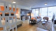 This thoughtfully designed single-floor space offers a seamless open-concept layout that integrates the sleeping, living, and kitchen areas into one cohesive, functional environment. Warm hardwood flooring flows throughout, tying the spaces together and adding a sense of warmth to the modern aesthetic.

The kitchen is well-appointed with dark espresso cabinetry, stainless steel appliances including a range, microwave, and dishwasher, complemented by a tile backsplash and dark countertops. A partial divider with decorative lattice detailing subtly separates the sleeping area, which features an accent wall in a bold orange hue, adding a vibrant pop of color and personality.

The living area is anchored by a plush sofa styled with orange accent pillows, a patterned area rug, and a dark coffee table, creating an inviting gathering space. The unit is flooded with natural light thanks to expansive floor-to-ceiling windows along the exterior wall, offering open views and a bright, airy ambiance. Exposed concrete ceilings contribute an industrial-modern character that balances the warmth of the wood flooring and the richness of the cabinetry, resulting in a stylish and contemporary feel throughout.