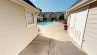 This outdoor amenity space centers around a large, crystal-clear swimming pool set within an expansive concrete deck designed to accommodate ample poolside lounging. The surrounding area is thoughtfully appointed with seating, including red lounge chairs and decorative potted flowers that add a welcoming touch of color to the environment.

The single-story pool house structure is finished with cream-colored horizontal lap siding and capped with a dark, pitched shingled roof, giving it a clean and traditional aesthetic. Multiple entry points — including classic six-panel doors and multi-pane French-style glass doors — provide convenient access to the interior facilities. A wall-mounted exterior light fixture ensures the area remains functional and safe into the evening hours.

The building's L-shaped layout creates a semi-enclosed courtyard feel around a portion of the deck, offering a sense of privacy while still opening up to the broader pool area. The surrounding community buildings, featuring multi-story residential architecture with balconies and traditional rooflines, are visible in the background and frame the space with a well-maintained neighborhood character. Under open skies with abundant natural light, this pool house and deck area offers a bright, inviting outdoor retreat suited for recreation and relaxation.