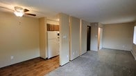 This single-floor residence offers a straightforward and functional layout designed for comfortable everyday living. The open-plan design allows for a seamless transition between the kitchen and the main living area, creating a sense of connected space throughout. The kitchen is equipped with warm wood-toned cabinetry, a full-size refrigerator, and a range, providing ample functionality for meal preparation. A textured ceiling runs throughout, adding a classic residential character to the interior. The living area features low-pile carpeting, while the kitchen zone transitions to hardwood-style flooring, clearly delineating the two spaces. Neutral beige walls create a clean, understated backdrop throughout the unit. Structural columns and partial walls add subtle architectural definition between areas without fully closing off the open feel. A hallway visible from the main living space leads to additional rooms, including what appears to be a bedroom or secondary living area with warm lighting, suggesting a private and comfortable retreat. Dark-finished interior doors provide a subtle contrast against the light walls. The overall ambiance is calm and practical, offering a welcoming environment suited to a variety of living arrangements.