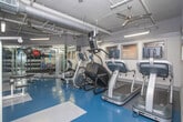 This fitness center is a fully equipped, single-level facility designed to support a wide range of workout routines. The space features a practical, no-frills aesthetic with gray walls, exposed ductwork along the ceiling, and bright overhead fluorescent lighting that keeps the environment well-lit throughout.

The flooring consists of durable rubberized tiles in a blue and gray checkered pattern, providing both safety and comfort underfoot. Floor-to-ceiling mirrors line one wall, creating a sense of expanded space while allowing users to monitor their form during exercise.

The cardio zone is lined with a StairMaster step mill, an upright stationary bike, and treadmills, all equipped with individual display screens. On the strength training side, Cybex International cable and weight machines are prominently featured, offering a professional-grade option for resistance training. A wall-mounted flat-screen TV provides entertainment near the strength equipment area.

A dedicated free weights section includes a fully stocked dumbbell rack, kettlebells in varying sizes, medicine balls, and a multi-functional RAX training rack with TRX suspension straps and resistance bands. An adjustable weight bench completes the free weight area. A vending machine is visible near the entrance, adding a convenient amenity for users. The facility offers a comprehensive and functional environment suitable for a full-body workout experience.
