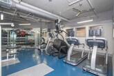 This fitness center is a fully equipped, single-level facility designed to support a wide range of workout routines. The space features a practical, no-frills aesthetic with gray walls, exposed ductwork along the ceiling, and bright overhead fluorescent lighting that keeps the environment well-lit throughout.

The flooring consists of durable rubberized tiles in a blue and gray checkered pattern, providing both safety and comfort underfoot. Floor-to-ceiling mirrors line one wall, creating a sense of expanded space while allowing users to monitor their form during exercise.

The cardio zone is lined with a StairMaster step mill, an upright stationary bike, and treadmills, all equipped with individual display screens. On the strength training side, Cybex International cable and weight machines are prominently featured, offering a professional-grade option for resistance training. A wall-mounted flat-screen TV provides entertainment near the strength equipment area.

A dedicated free weights section includes a fully stocked dumbbell rack, kettlebells in varying sizes, medicine balls, and a multi-functional RAX training rack with TRX suspension straps and resistance bands. An adjustable weight bench completes the free weight area. A vending machine is visible near the entrance, adding a convenient amenity for users. The facility offers a comprehensive and functional environment suitable for a full-body workout experience.