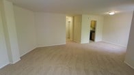 This single-floor residential space offers a clean, functional layout designed for comfortable everyday living. The main living area is notably open and spacious, finished with wall-to-wall neutral-toned carpeting that adds warmth and softness underfoot. The walls are painted in a light, neutral tone throughout, contributing to a bright and airy atmosphere that makes the space feel inviting.

A central hallway extends from the living area, providing access to additional rooms within the unit, including what appears to be a bathroom or utility closet with a door visible at the end. To the right of the living area, an open doorway leads directly into the kitchen, which features cabinetry in warm wood tones, dark countertops, and dark appliances. The kitchen is compact yet well-organized, offering the essential amenities for everyday meal preparation.

White baseboard trim runs along the perimeter of the living area, providing a crisp contrast to the carpeted floors and light-colored walls. The overall layout is straightforward and efficient, with a seamless flow between the main living space, kitchen, and the rest of the unit. The design prioritizes practicality while maintaining a neutral aesthetic that lends itself well to a variety of interior styles.