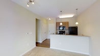 This single-floor unit offers a thoughtfully designed open-concept layout that connects the living area to the kitchen, creating a spacious and functional environment. The living area features neutral carpet flooring and track lighting, contributing to a bright and welcoming atmosphere.

The kitchen is a standout feature, equipped with black appliances including a refrigerator, range, and microwave. Warm wood-toned cabinetry provides ample storage, while granite-look countertops add a polished finish. A peninsula counter separates the kitchen from the living space, offering additional prep space and casual seating potential. Pendant lighting above the peninsula enhances both the aesthetic and functionality of the kitchen.

Durable hardwood-style flooring in the entryway and hallway provides a natural transition between rooms. The hallway leads to the bedroom and bathroom, offering a practical layout with clear separation between living and private spaces. Neutral wall tones throughout the unit create a clean, versatile canvas that complements a variety of interior styles.