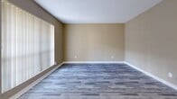 Located at 10501 Holly Springs Dr in Houston, Texas, this single-story home offers a clean, updated interior with a cohesive design throughout. The flooring is a standout feature, with grey wood-look planks extending consistently across all living areas, bedrooms, and hallways, lending a modern and unified aesthetic to the space.

The living area is generously proportioned, featuring warm beige walls, crisp white baseboards, and large windows dressed with vertical blinds that allow ample natural light to fill the room. A hallway connects the living areas to the private bedrooms, each of which is equipped with a ceiling fan and light fixture, vertical-blind windows, and the same durable plank flooring found throughout the home.

One bedroom features access to a walk-in closet and an en-suite bathroom, which includes a granite-topped vanity with white cabinetry, a tiled shower-tub combination, and matching plank flooring. The kitchen area showcases granite countertops, white cabinetry, and includes built-in appliances. A door from the kitchen provides additional access within the layout.

The overall ambiance of this home is clean, neutral, and move-in ready, with consistent finishes and practical details that make it a well-maintained and functional single-floor residence.