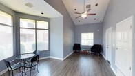 This single-floor clubhouse offers a well-designed communal space that balances comfort and functionality. The main gathering area immediately impresses with its vaulted ceilings, which lend a sense of openness and grandeur. A ceiling fan is centrally mounted, ensuring year-round comfort throughout the space. The floors are finished in a warm hardwood-style material that adds a polished, modern aesthetic and is easy to maintain. Walls are painted in a cool, neutral gray tone that creates a calm and inviting atmosphere throughout.

Natural light is a defining feature of this clubhouse, with multiple large windows fitted with horizontal blinds allowing residents to control the amount of light and privacy as needed. Views of the surrounding outdoor environment — including glimpses of neighboring buildings and greenery — can be enjoyed from multiple vantage points within the space.

The seating arrangement includes a pair of dark accent chairs positioned around a glass-top coffee table, creating an intimate conversation area near one of the windows. A small bistro-style table and chairs are positioned near the large front-facing window, offering a cozy spot for casual dining or socializing. Recessed lighting complements the natural light, ensuring the space remains bright and functional at all hours. Overall, the clubhouse provides a versatile and attractive environment for community gatherings.
