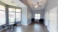 This single-floor clubhouse offers a well-designed communal space that balances comfort and functionality. The main gathering area immediately impresses with its vaulted ceilings, which lend a sense of openness and grandeur. A ceiling fan is centrally mounted, ensuring year-round comfort throughout the space. The floors are finished in a warm hardwood-style material that adds a polished, modern aesthetic and is easy to maintain. Walls are painted in a cool, neutral gray tone that creates a calm and inviting atmosphere throughout.

Natural light is a defining feature of this clubhouse, with multiple large windows fitted with horizontal blinds allowing residents to control the amount of light and privacy as needed. Views of the surrounding outdoor environment — including glimpses of neighboring buildings and greenery — can be enjoyed from multiple vantage points within the space.

The seating arrangement includes a pair of dark accent chairs positioned around a glass-top coffee table, creating an intimate conversation area near one of the windows. A small bistro-style table and chairs are positioned near the large front-facing window, offering a cozy spot for casual dining or socializing. Recessed lighting complements the natural light, ensuring the space remains bright and functional at all hours. Overall, the clubhouse provides a versatile and attractive environment for community gatherings.